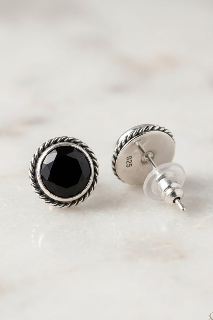 A pair of sterling silver stud earrings with a faceted black onyx center and a twisted rope border, shown on a light marble surface. One earring is facing forward, and the other is turned to show the post and backing.