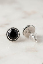 A pair of sterling silver stud earrings with a faceted black onyx center and a twisted rope border, shown on a light marble surface. One earring is facing forward, and the other is turned to show the post and backing.