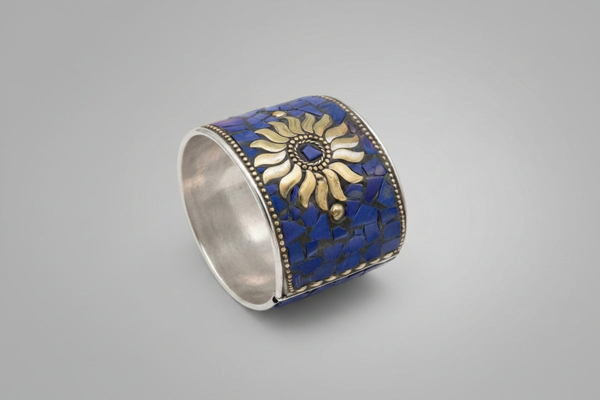 Sterling silver cuff bracelet with a blue lapis lazuli mosaic exterior and a gold sunburst motif, shown with a product tag on a neutral gray background.