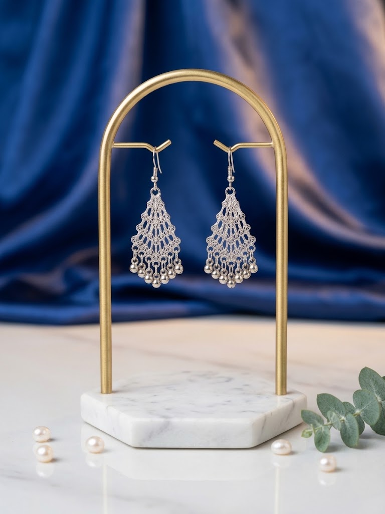 A pair of silver-toned dangle earrings with intricate filigree and pearl bead details, displayed on a gold-framed stand over a white marble base. The setup is styled with scattered pearls and green leaves against a draped blue fabric background.