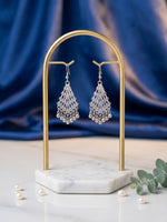 A pair of silver-toned dangle earrings with intricate filigree and pearl bead details, displayed on a gold-framed stand over a white marble base. The setup is styled with scattered pearls and green leaves against a draped blue fabric background.