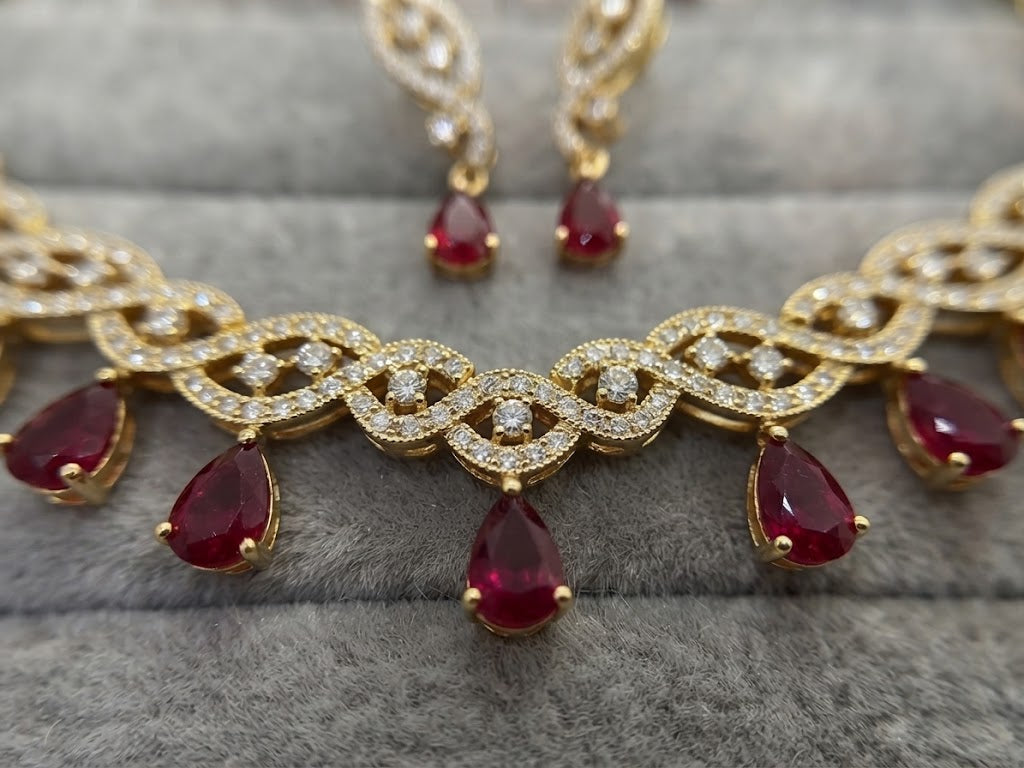 Gold-toned traditional Indian jewelry set featuring a necklace and matching earrings with teardrop-shaped red gemstones and pavé-set clear stones, displayed on a gray textured velvet surface.