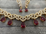 Gold-toned traditional Indian jewelry set featuring a necklace and matching earrings with teardrop-shaped red gemstones and pavé-set clear stones, displayed on a gray textured velvet surface.