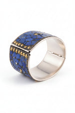 A wide silver-toned cuff bracelet with a textured blue mosaic inlay and rows of small gold studs, photographed on a dark fabric background.