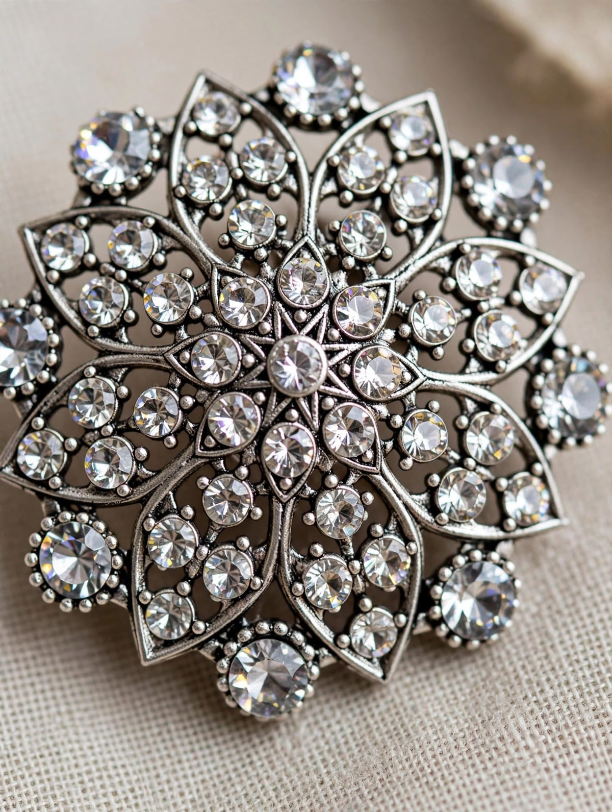 A close-up of a silver-toned brooch featuring an intricate floral or snowflake-like design set with clear rhinestones, resting on a textured beige fabric background.