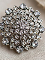 A close-up of a silver-toned brooch featuring an intricate floral or snowflake-like design set with clear rhinestones, resting on a textured beige fabric background.