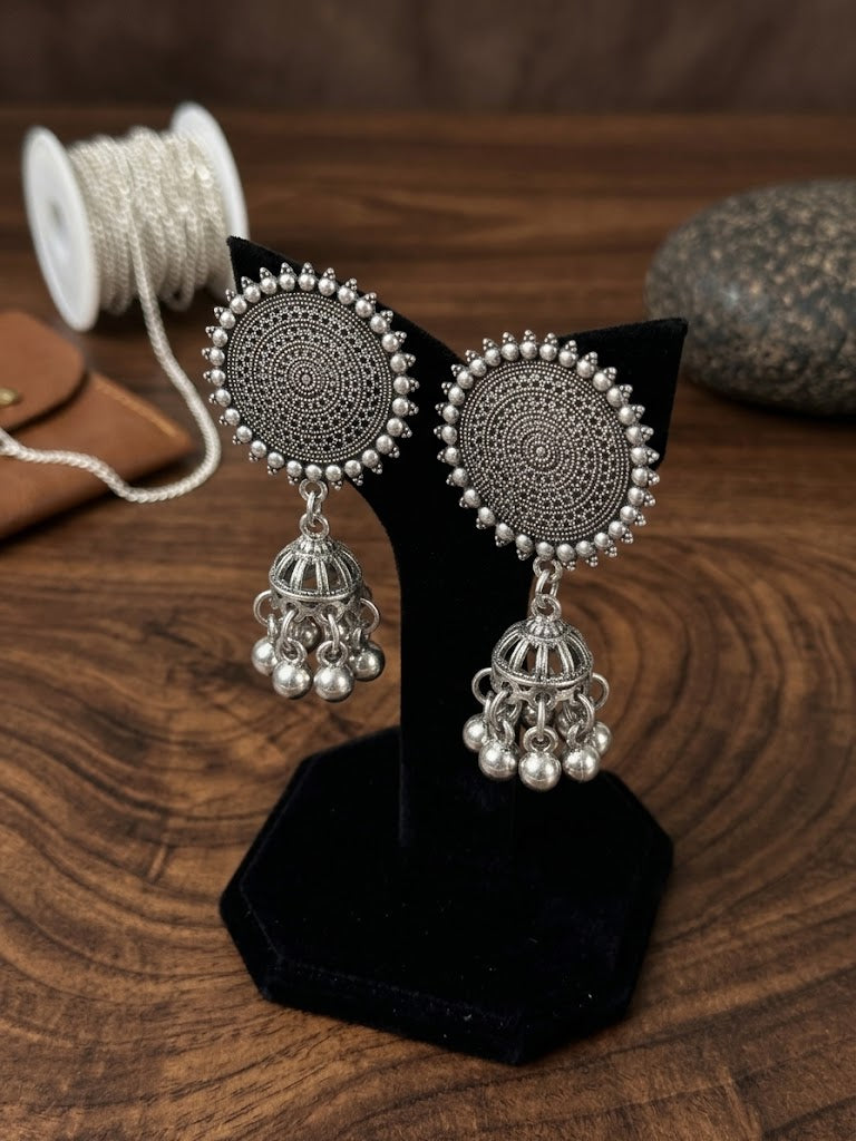 A pair of silver-toned Indian jhumka earrings with intricate circular filigree patterns and dangling bell charms, displayed on a black stand over a wooden surface.