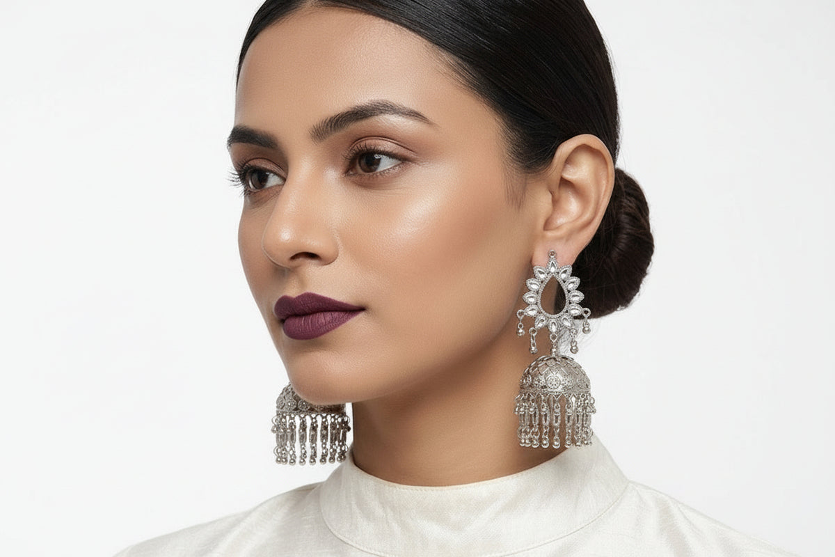 A close-up of a woman wearing ornate silver-toned Indian jhumka earrings with dangling tassel chains, paired with a white high-neck top against a light gray background.