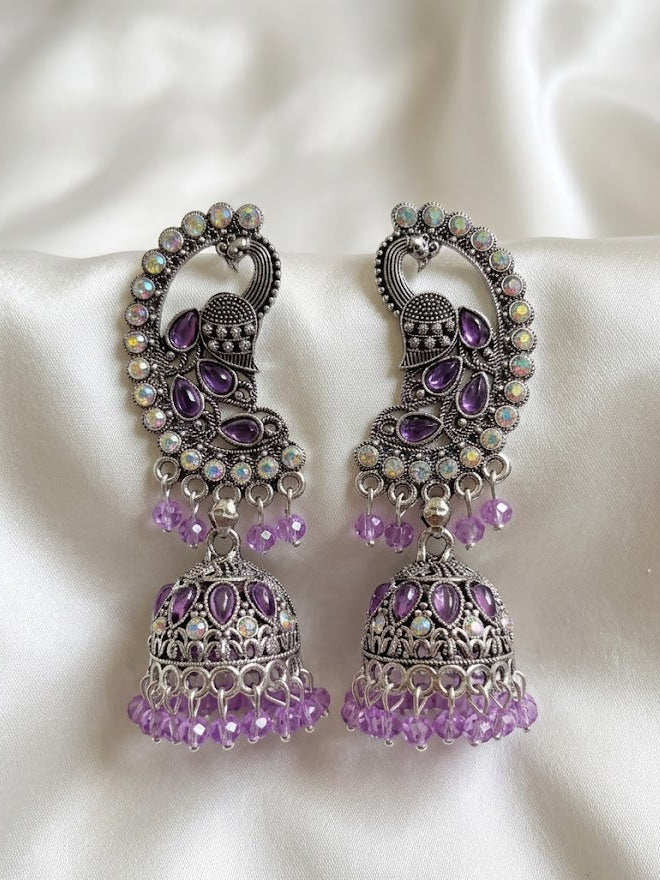 A pair of ornate silver-toned Jhumka earrings decorated with purple amethyst stones and iridescent rhinestones, resting on a white satin fabric background.