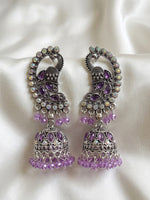A pair of ornate silver-toned Jhumka earrings decorated with purple amethyst stones and iridescent rhinestones, resting on a white satin fabric background.