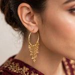 A close-up of a woman's ear wearing a gold huggie hoop earring with dangling beaded tassels, paired with a deep maroon embroidered garment.