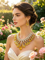 A woman in a white strapless dress wearing a dramatic gold statement necklace and matching drop earrings with large yellow teardrop gemstones, photographed outdoors in a sunlit garden with pink flowers.