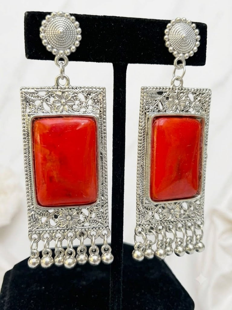 A pair of silver-toned statement earrings with large rectangular red stone centers and dangling silver filigree and bead fringe, displayed on a black jewelry stand against a light fabric background.