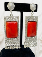 A pair of silver-toned statement earrings with large rectangular red stone centers and dangling silver filigree and bead fringe, displayed on a black jewelry stand against a light fabric background.