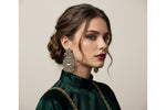 A woman with styled brown hair in an updo wearing a dark green velvet high-neck top with gold embroidery, showcasing a large, ornate silver-toned traditional Indian jhumka earring.