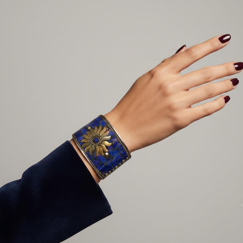 A close-up of a person's hand wearing a wide blue cuff bracelet with a gold sunburst design and dark blue nail polish, set against a blurred background of antique bookshelves.