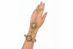 An ornate gold-toned Indian style manjika bracelet with matching ring and a wrist chain, decorated with gold filigree, white stones, and a central teardrop-shaped crystal, shown on a hand against a white background.