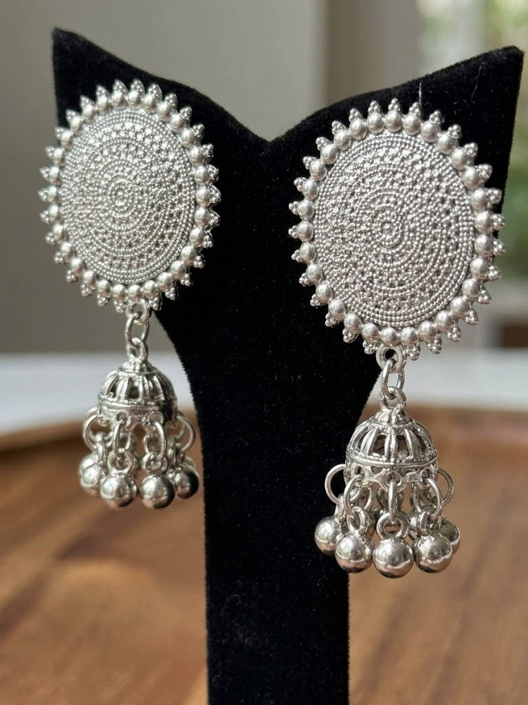 A pair of intricate silver-toned Indian jhumka earrings displayed on a black velvet stand. The earrings feature circular, heavily textured mandala designs with dangling bell-shaped charms.