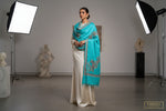 The Turquoise Kashmiri Aari embroidered marina scarf with tassels hanging at the ends, displayed on a wooden hanger with greenery in the background.