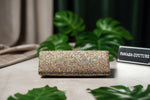 The clutch bag with a light base covered in multi-colored nano glass beads, displayed on a wooden surface surrounded by green leaves.