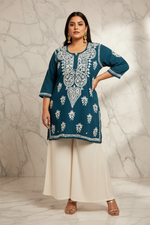 A woman models a teal blue kurta with intricate white embroidery, paired with wide-leg cream trousers and gold sandals, standing against a marble-patterned wall.