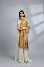 The mustard Kashmiri Aari embroidered marina scarf with tassels hanging at the ends, displayed on a wooden hanger with greenery in the background.