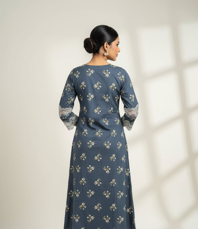 The rear view of a state blue long-sleeve kurta with a beige floral pattern and white lace trim on the cuffs and hem, laid flat on a light wood floor.