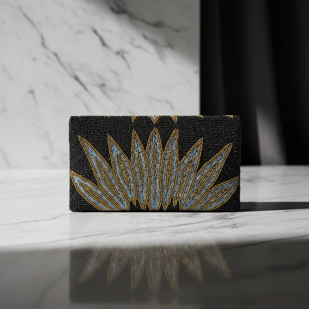 The rear view of this handcrafted clutch bag with a geometric pattern of glass beads in gold and blue on a black base, displayed on a wooden surface with green leaf accents around.