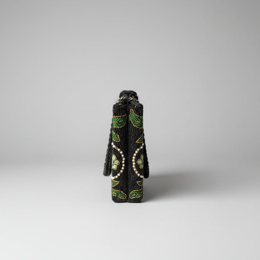 The side view of a black floral handcrafted tote bag with glass beads embroidered on a white silk base, displayed on a wooden surface with a greenery background