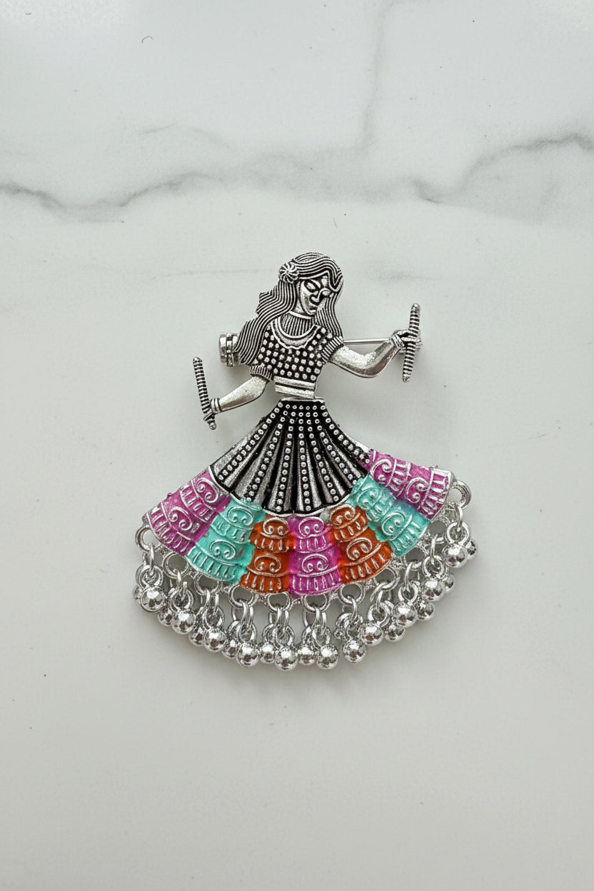 The silver-toned dholak dancer earrings with multi-coloured beaded skirts, displayed on a white marble surface with colourful decorative objects in the background.