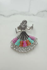 The silver-toned dholak dancer earrings with multi-coloured beaded skirts, displayed on a white marble surface with colourful decorative objects in the background.
