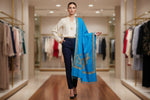 The sky blue Kashmiri Aari embroidered marina scarf with tassels hanging at the ends, displayed on a wooden hanger with greenery in the background.