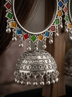 A close-up of a silver-toned traditional Indian jhumka earring with colorful square gemstones in red, green, blue, and orange, hanging from a silver-toned circular band. The earring is shown worn on a person's ear with dark hair visible, against a warm brown background.