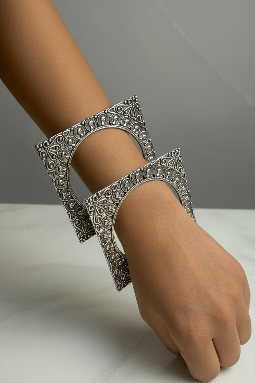 Two ornate silver-toned bangles with intricate filigree patterns and small clear stones, worn on a person's wrist against a light marble-like background.