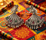 Two ornate silver-toned jhumka earrings shaped like dancing figures, decorated with green and red stones and dangling bells, displayed on a colorful embroidered textile with wooden sticks.