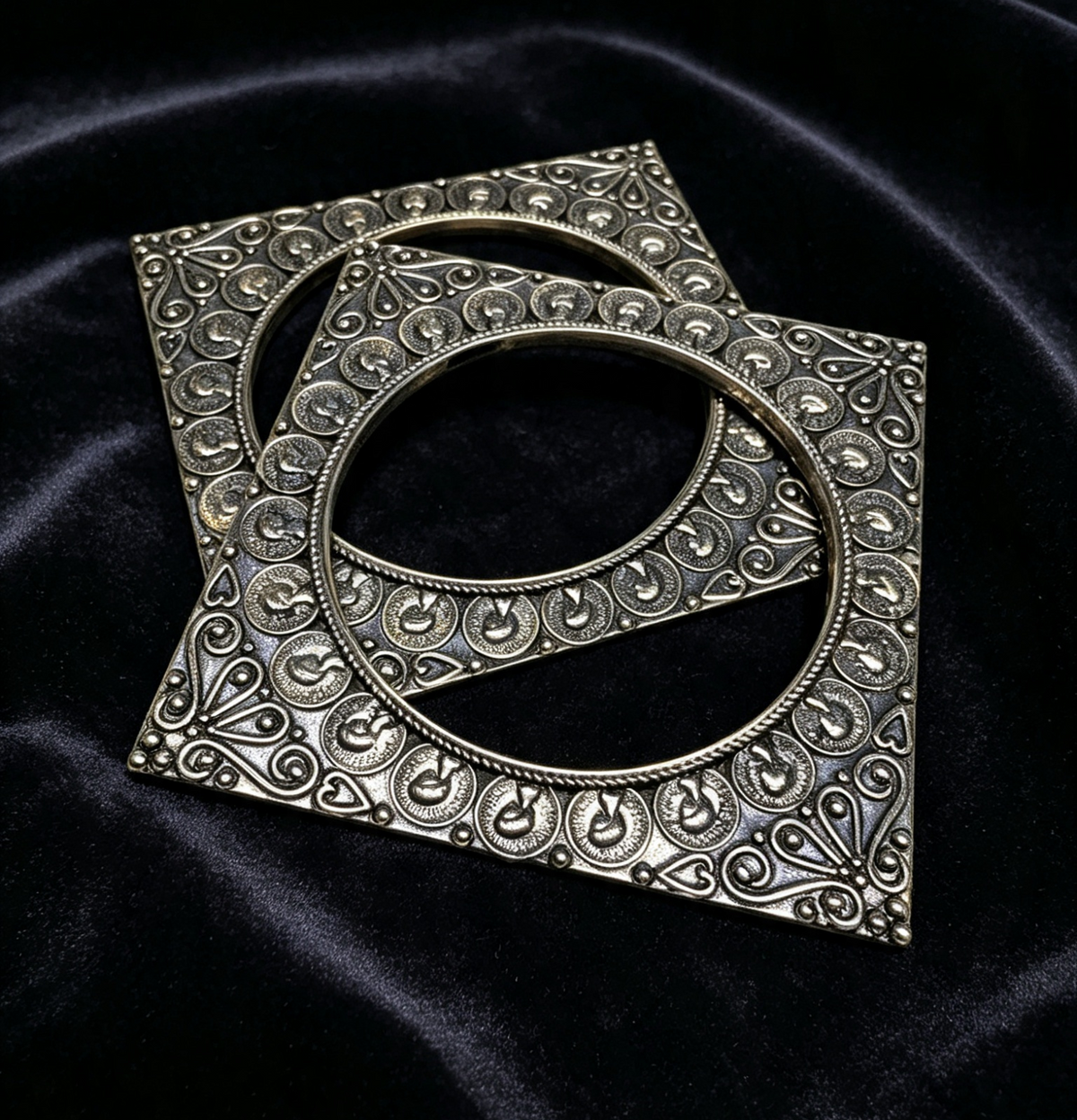Two ornate silver-toned square metal frames with circular cut-outs and intricate filigree detailing, displayed on a dark black velvet fabric background.