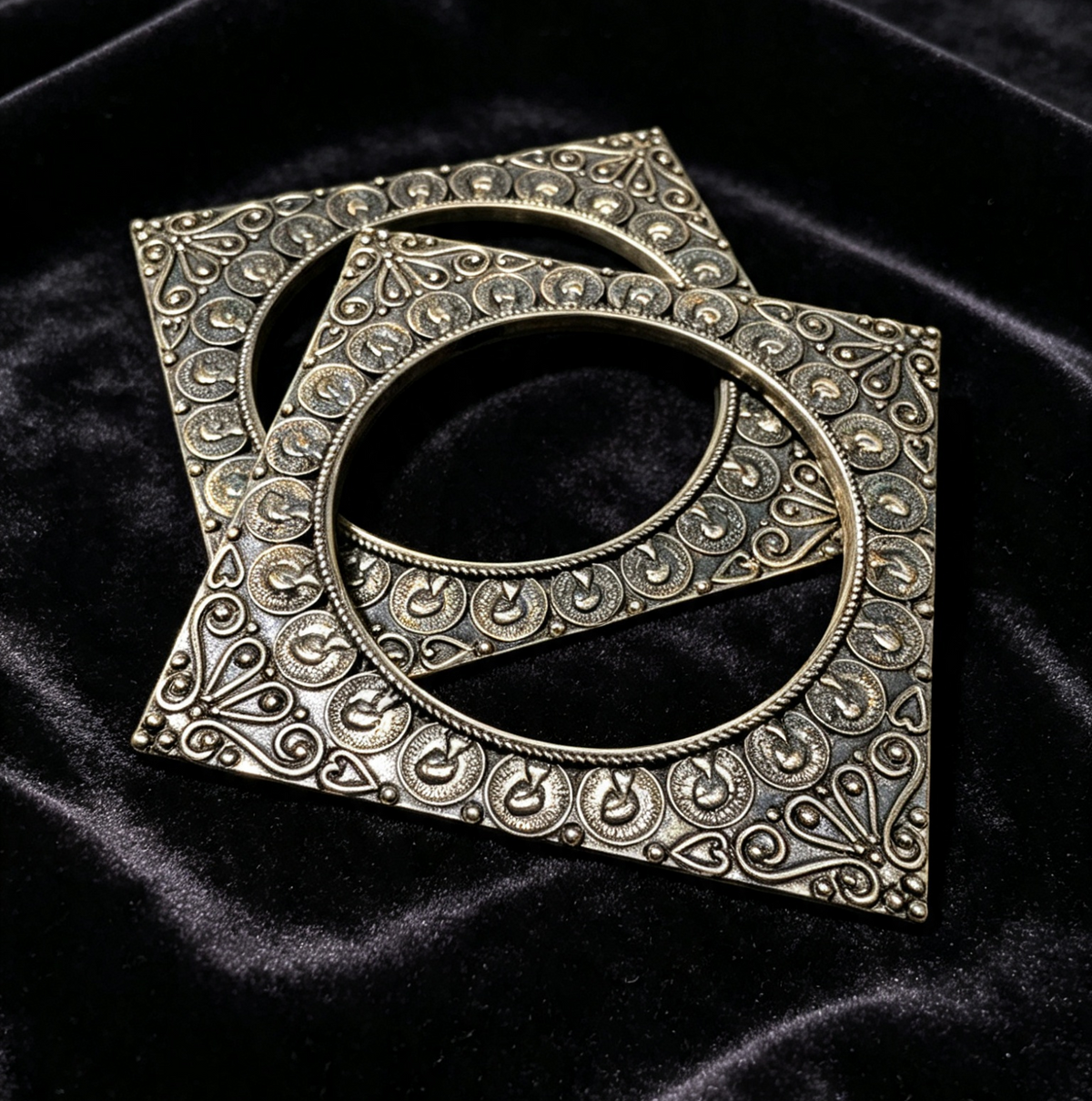 Two square metallic oxidised slide on bangles frames with intricate filigree detailing, displayed on a black velvet fabric background.