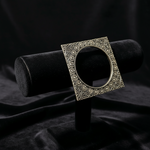 An ornate square silver-toned ring box with a decorative filigree border, displayed on a black velvet jewelry stand against a dark draped fabric background.