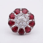 A silver-tone ring featuring a circular arrangement of deep red teardrop-cut gemstones surrounding a central cluster of clear marquise and round-cut stones, set against a plain white background.