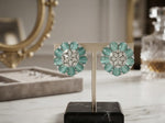 A pair of floral cluster earrings displayed on a stand on a marble surface. The earrings feature mint green marquise-cut stones and clear round stones in a silver-toned setting, with a blurred luxury background including a gold mirror frame and a crystal decanter.