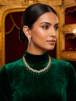 A woman in a dark green velvet turtleneck wears a delicate gold chain necklace with green stone accents and matching drop earrings, posed in front of an ornate gold and red interior.
