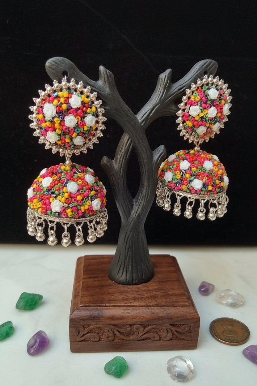 A pair of multicolor beaded Indian jhumka earrings displayed on a dark tree-shaped jewelry stand. The earrings feature intricate silver-toned filigree borders, colorful seed bead patterns in pink, yellow, orange, green, and white, and dangling silver-toned bell charms.