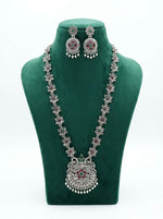 Long oxidized silver necklace and matching earrings with floral patterns and pink and green stones displayed on a green velvet stand.