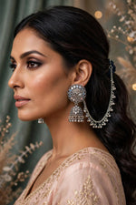 A woman in profile wearing ornate silver-toned jhumka earrings with dangling chains and a decorative circular top, paired with a light beige embroidered garment. The background is softly blurred with warm bokeh lights and neutral drapery.