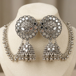 Silver-toned Indian jhumka earrings and matching necklace displayed on a cream-colored jewelry bust. The intricate filigree features circular mirror work and dangling bell-shaped jhumkas.