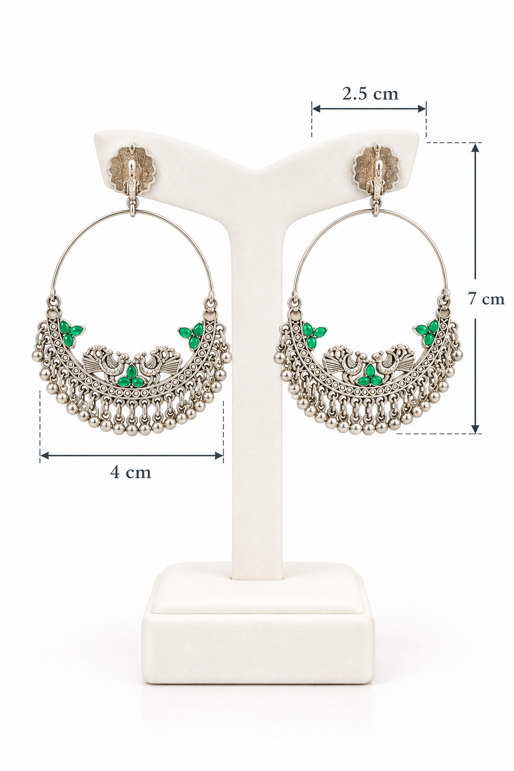 A pair of oxidized silver hoop earrings featuring irregular textured studs, traditional peacock motifs with vibrant green stone accents, and hanging bead tassels, displayed on a black stand