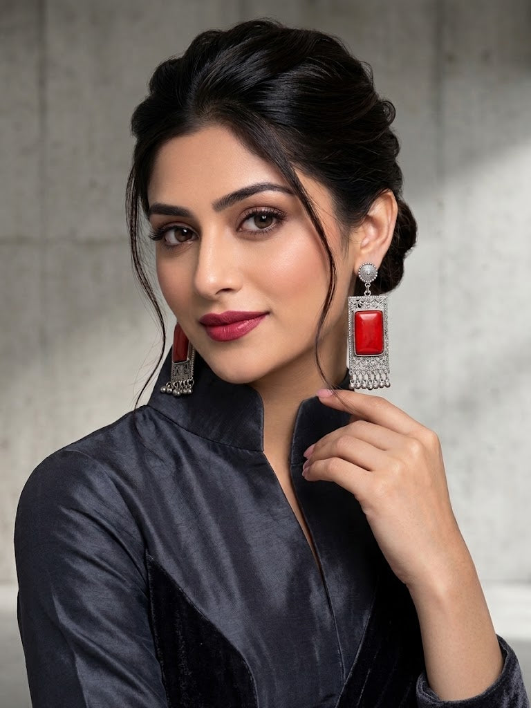 A woman with dark hair styled in an updo wears a black satin top and large silver-toned statement earrings with red rectangular inlays, posing against a textured gray concrete wall.