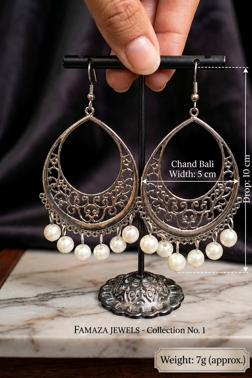 A pair of ornate silver-toned Chand Bali earrings with dangling white pearls, displayed on a black stand with a hand holding the top bar. White dimension lines show the drop of 10 cm, width of 5 cm, and the earrings are labeled fAMAZA JEWELS Collection No.