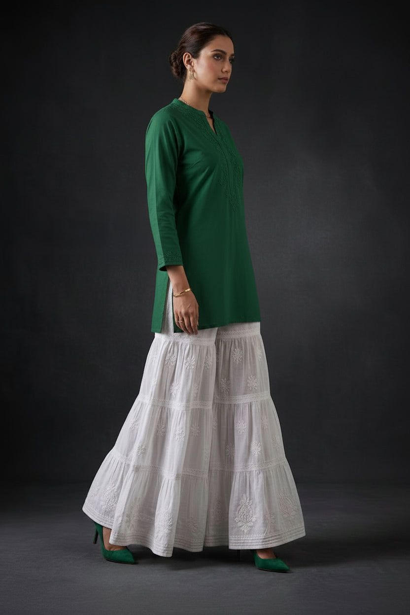 A model standing sideways wearing a white 4-tier cotton flared sharara with intricate white Chikankari embroidery, posed against a dark background.