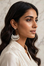 Woman wearing luminous all-silver Chandbali hoop earrings with clear stone inlay and cascading round white pearls, styled in cream embroidered outfit against soft marble background — timeless bridal & formal jewelry.